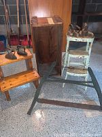 Photo showing the large antique bow cross cut saw, wooden stool, wooden crate, heavy wooden steps, and five antique irons arranged on concrete floor.