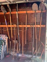 Long-handled garden tools hanging on a wooden wall including shovels, rakes, forks, and hoes