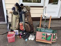 Photo showing three golf bags filled with various golf clubs, indoor putting green set, golf shoes, golf balls, and paraphernalia arranged on ground outside a building.