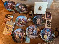 Photo showing all seven Hummel plates, three figurines, two books, a small bell, and an ornament on a wooden floor