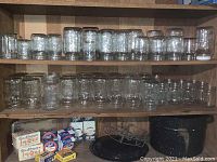 Wide view of kitchen shelf showing multiple rows of various sizes and types of clear glass mason jars, some with embossed branding. Boxes of Gulfwax and a black speckled enamel canning pot with rack on lower shelf.