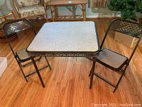 Overall view of square children's folding table with two matching folding chairs in black perforated metal and laminate top on wood floor.