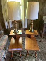 Two mid century two-tier wooden end tables each topped with a wooden based lamp with a fabric shade in a living room setting.