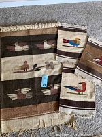 Four woven textiles laid on floor, showcasing southwestern style bird patterns in earthy tones with fringed edges. Larger textile includes a hanging wood rod.