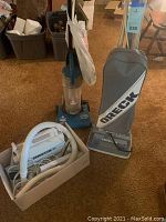 Two Oreck vacuum cleaners (one canister and one upright) and one Bissell upright vacuum in a room with brown carpet, some boxes in background.