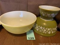 Three Pyrex mixing bowls shown together. Two green bowls with white Spring Blossom floral pattern stacked, next to a solid yellow bowl. Displays size difference and condition.
