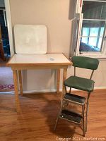 View of the tall kitchen table with white surface, wood edges, extra board leaning on the wall and the green vinyl stool next to it