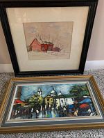 Photo showing two framed paintings leaning against a wall: one a red barn in winter in a dark frame, the other a colorful cityscape with people and umbrellas in a gold and gray frame.