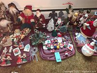 Wide view showing the majority of Christmas decorations arranged on folding tables and carpet floor. Includes Santa dolls, nutcrackers, ornaments, cardboard figures, wreaths, and small collectibles.