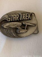Front view of oval metal belt buckle with raised Star Trek logo and USS Enterprise starship design in antiqued finish.