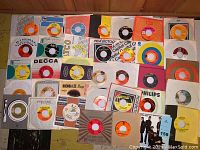 Top view of over 35 vintage 45 RPM vinyl records laid out showing various colorful labels and sleeves including RCA Victor, Decca, Philips, and more.