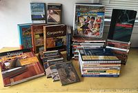 Wide view of a large collection of hardback and softcover books and DVDs on carpentry and construction topics arranged on a table, showing titles and various instructional materials.