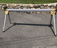 Full view of DEWALT miter saw stand fully assembled outdoors on asphalt with stone wall background.