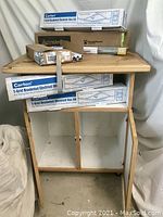 Rolling wooden cabinet with natural wood top and white doors, showing wear and in fair condition, with boxed electrical and drawer slide kits placed on top and inside the open shelf area.