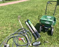 Full lot laid on grass: Scott's Turfbuilder Mini spreader, gardening hoe, two rolls of barrier netting, garden hose with nozzle and sprinkler attachments.