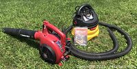 Troy-Bilt TB430 red gas leaf blower with black blower tube alongside yellow and black Stinger wet/dry vacuum with attached hose on grass.