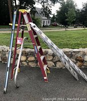 Red Werner 6 ft. step ladder, aluminum extension ladder and loading ramp outdoors on pavement with stone wall background.