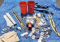 Overview of the painting and drywall tools displayed on blue tarp including brushes, rollers, buckets, drywall knives, hawk, and suspenders.