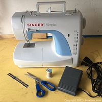 Full side view of Singer Simple sewing machine with foot pedal, spool of thread, scissors, and measuring tool on yellow work surface.