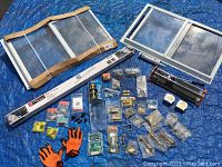 Photo showing two white sliding three-track storm windows, a long LED light strip box, two caulk guns, small plastic bags with assorted brass hardware, tape measures, orange gloves, and other various tools and small hardware parts arranged on a blue tarp.