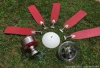 Disassembled five red blades, brushed nickel motor housing with red accents, frosted white glass light cover, and fan wheel laid out on grass.