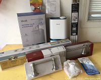 Full lot of home improvement items including the Levoit humidifier with its box and manual, bathroom light bar, towel bar, toilet paper holder, glass doorknob set, chain doorstop, lamp base and screen kit, all on a table.