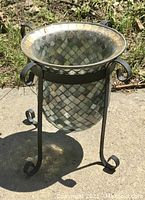 Decorative deep stained glass style planter in black wrought iron scroll base, placed on concrete with grass around.