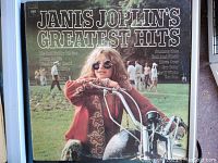 Front cover of Janis Joplin's Greatest Hits 1973 vinyl album showing Janis on a motorcycle with background figures and track list.