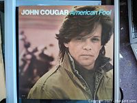 Close-up photo of John Cougar American Fool album cover showing the artist in a green jacket with text title and artist name.