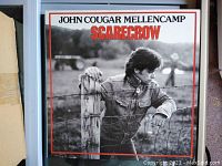 Front cover of John Cougar Mellencamp's 1985 Scarecrow vinyl LP record showing the black-and-white photo of the artist by a fence.
