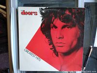 Close-up image of the vinyl record album cover showing Jim Morrison's face in red and white color scheme with The Doors band name and Greatest Hits title.