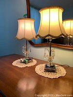 Pair of glass and crystal prism bedside lamps on a wooden table with lace doilies, turned on, showing fabric shades with fringed trim and clear glass bases with hanging prism details.