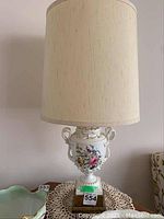 Full view of ceramic lamp showing fabric shade and floral china base on table with crochet doily.