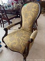 Front angled view showing carved wooden frame and floral patterned upholstery on backrest and seat, padded armrests, and castor feet.