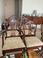 Five wooden side chairs and one captain's chair with upholstered seats and pierced splat back design, arranged in a room with other furniture and tableware in the background.