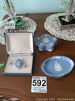 Photo showing the entire set: boxed brooch and earrings, trinket box, two small dishes on a wooden table with lace doilies and decorative lamp bases in the background.
