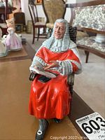 Seated porcelain figure of a judge in a red robe and white wig, holding an open book, frontal view.