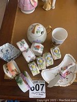 Photo shows ceramic napkin rings with various floral and scenic designs, a Royal Crown Derby cheese plate and spreader, mini serving plates, a mini pot, and a small cup on table.