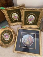 Four framed painted porcelain plaques displayed on floor showing detail of ornate gold frames and painted porcelain scenes.