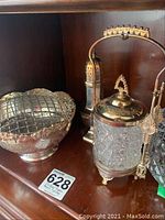 Shelf display showing three silverplate items: a bowl with flower frog insert on the left, pickle jar with tongs on the right, and sugar caster in the back center.