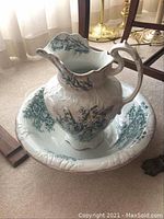 Wide view showing the antique water pitcher inside matching ceramic basin on carpeted floor with brass lamps in background