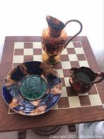 Three items displayed on a wooden chessboard table: a Wedgwood plate, copper lustreware jug, and copper ewer.