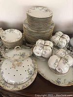 Overview of the stacked plates, tea cups, vegetable dish with lids, and serving platter displaying floral violet pattern with gold trim.
