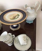 Full view of cake stand, antique jug, and some demitasse cups and saucers on table.