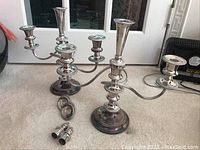 Pair of three-arm silverplate candelabra with central taller candle holder, sitting on carpet. Includes candle snuffers and drip catchers in front.