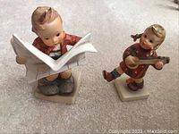 Two Hummel porcelain figurines standing on a light grey carpeted surface: a boy reading a newspaper on the left and a girl playing a guitar on the right.