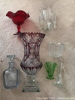 Seven items arranged on carpet: red compote, clear pitcher, amethyst vase, green cordial, decanter, flared bowl, fluted vase