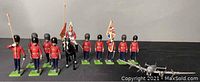 Eleven red-uniformed Scots Guards toy soldiers arranged with one soldier mounted on black horse holding a red banner and an airplane figure in front.