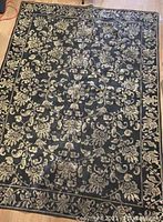 Full view of the black Persian-style rug with beige and taupe floral and vine motifs laid flat, showing minor wrinkles and wear.