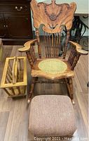 Hand carved wooden rocking chair with detailed floral and scroll carving on backrest, caned seat, wooden arms and spindles, positioned with foot stool and magazine rack